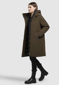 Oliven grøn, knælang parka med hætte, sort lynlås og to sidelommer. Har en slank, glat tekstur og minimalistisk design.
