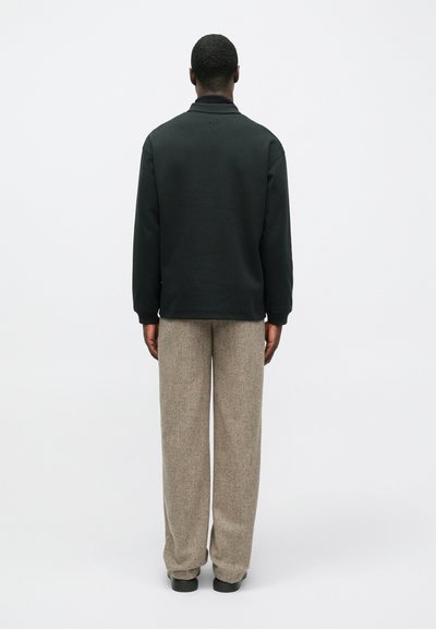 Άνδρας που στέκεται με την πλάτη γυρισμένη, φορώντας ένα μαύρο μπλουζάκι με μακριά μανίκια και χαλαρούς μπεζ παντελόνες, μπροστά σε λευκό φόντο.