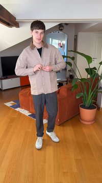 Beige Hemdjacket mit zwei Taschen, getragen über einem schwarzen Hemd. Graue strukturierte Hose und weiße Sneakers. Innenraum mit einer Pflanze.