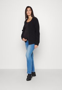 Cardigan negro de punto, con botones en la parte delantera, llevado sobre una camiseta negra, combinado con jeans de campana azules y zapatos negros. Textura suave; ajuste casual.