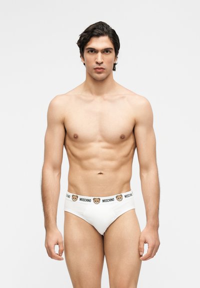 Άνδρας μοντέλο χωρίς πουκάμισο με σκούρα μαλλιά, φοράει λευκά εσώρουχα Moschino με προσωπάκια αρκούδων στη μέση, στέκεται μπροστά σε απλό φόντο.