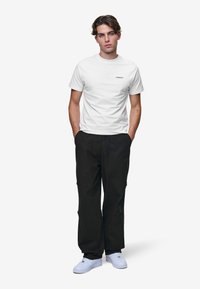 Ung man som står med händerna i fickorna, klädd i en vit t-shirt, svarta byxor, vita sneakers och en liten tatuering på armen.