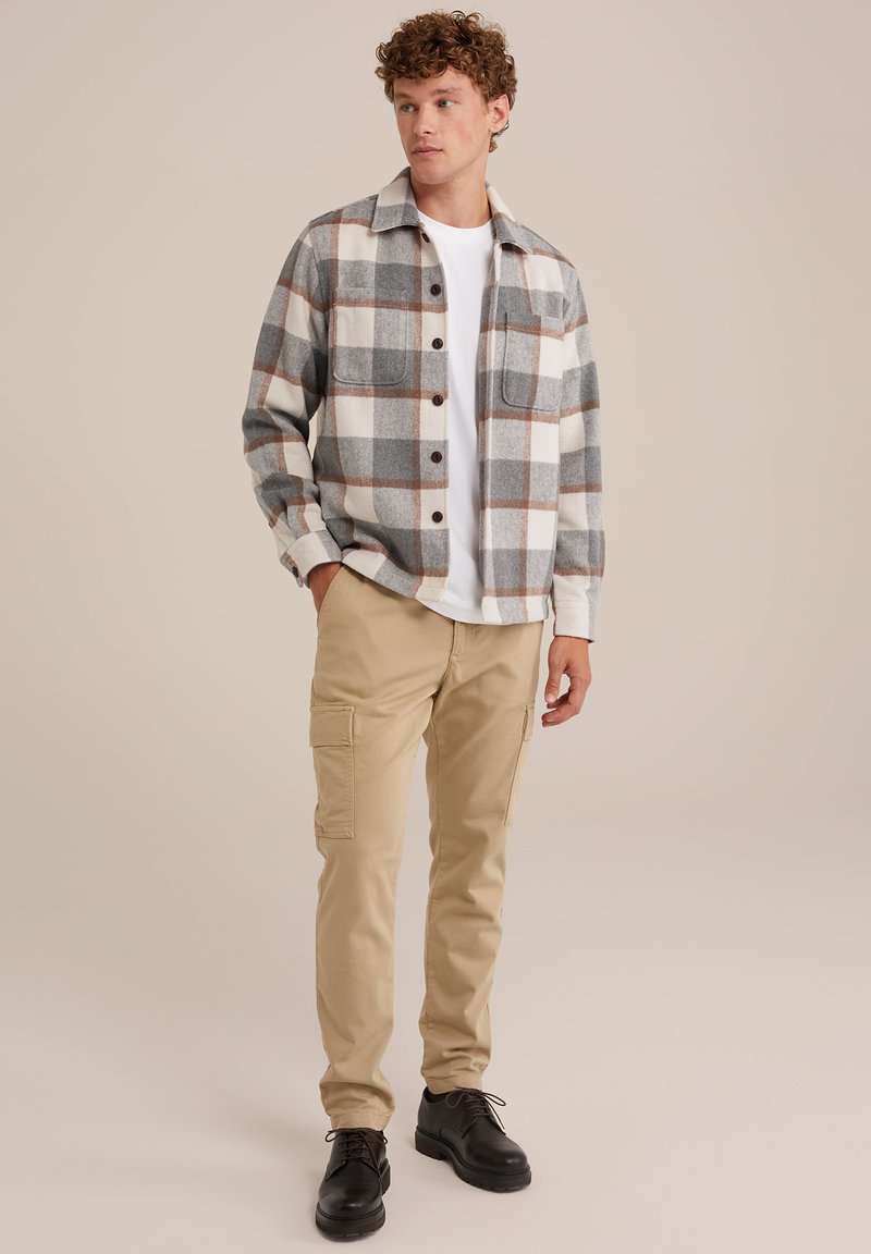 Młody mężczyzna stoi z rękami w kieszeniach, ubrany w beżowe spodnie cargo, białą koszulkę, szaro-brązową koszulę w kratę i czarne sznurowane buty na jednolitym tle.