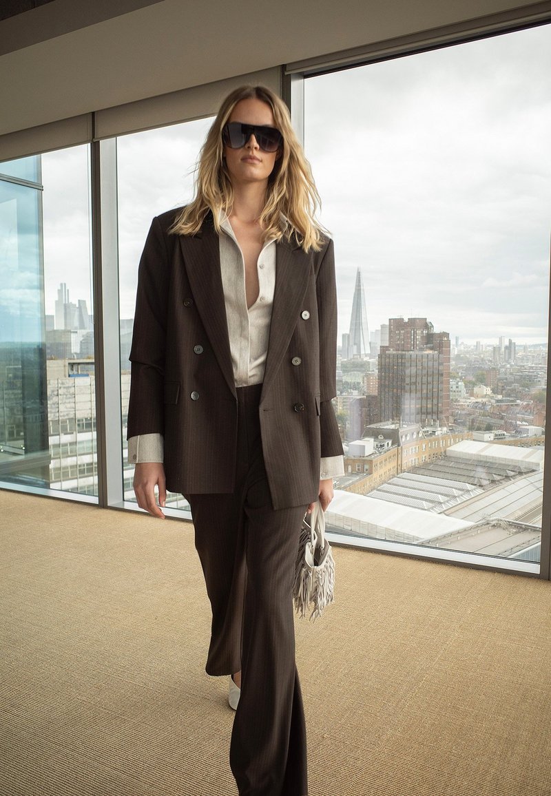 Femme en tailleur marron foncé et chemise blanche, portant des lunettes de soleil et tenant un sac à franges, marchant à l'intérieur près d'une grande fenêtre avec vue sur la ville.