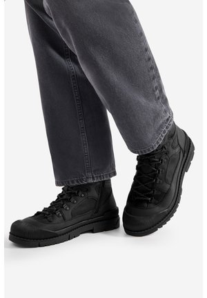 Czarne buty trekkingowe z teksturowanymi gumowymi podeszwami, wiązane na sznurowadła, z wzmocnionymi noskami, zestawione z szarymi, prostymi jeansami.