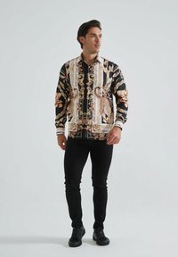 Chemise à manches longues noire avec un motif baroque floral et doré, dotée d'un col, d'une fermeture éclair et associée à un jean slim noir et des baskets.