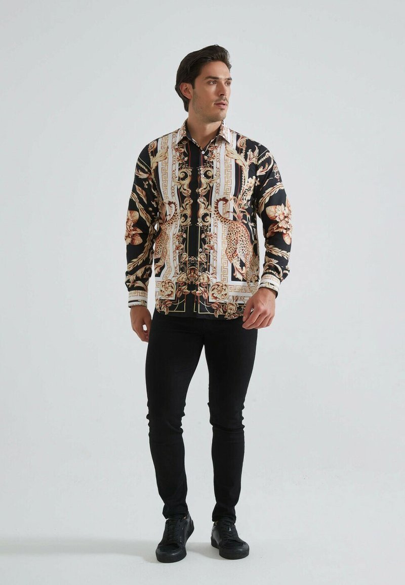 Chemise à manches longues noire avec un motif baroque floral et doré, dotée d'un col, d'une fermeture éclair et associée à un jean slim noir et des baskets.