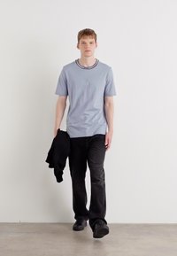 Lyseblå kortærmet t-shirt med en sort ribbestrikket krave, parret med mørke bukser. Modellen holder en sort jakke og har sorte sko på.