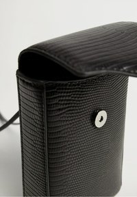 Černá texturovaná kožená taška s hladkým povrchem, magnetickým uzávěrem a štíhlým designem. Obsahuje úzký ramenní popruh.