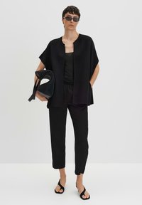 Veste cape oversize noire sur un haut noir, associée à un pantalon noir fuselé et des sandales flip-flop. Tient un sac à main noir texturé.