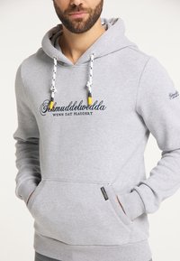Homme portant un sweat à capuche gris clair avec des cordons blancs et un texte en script noir "Schmuddelwedda WENN DAT PLADDERT" sur le devant, les mains dans les poches.