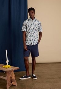 Korte mouwen, lichtkleurig shirt met bloemenpatroon, gecombineerd met donkerblauwe shorts en marineblauwe sneakers op een eenvoudige houten tafel.