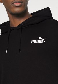 Personne portant un sweat à capuche noir avec un logo Puma blanc sur la poitrine gauche et des cordons visibles.