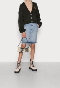 Persona che indossa un cardigan nero con bottoni sopra un dolcevita beige, una gonna di jeans e stivali beige con lacci, mentre tiene in mano una borsa con motivo in pelle di serpente.