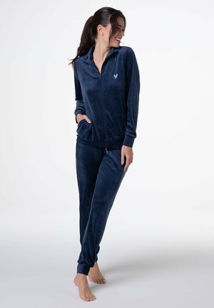 Donna sorridente, che indossa una giacca zip-up in velluto blu navy e pantaloni coordinati, in piedi scalza contro uno sfondo bianco.