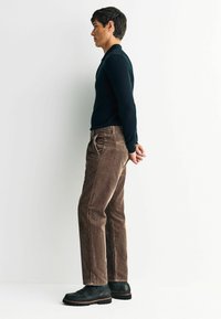 Brown corduroy pants with a straight-cut design, paired with a fitted black long-sleeve shirt and black footwear. Side view.