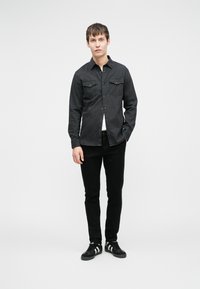 Giovane in piedi, indossa una camicia nera con bottoni, pantaloni neri e sneakers nere con strisce bianche, di fronte a uno sfondo bianco uniforme.