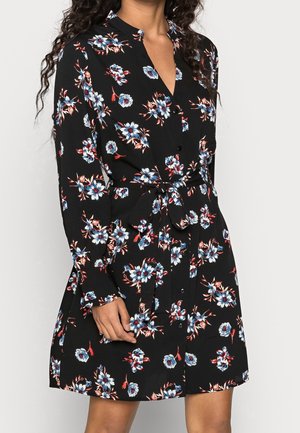 Kvinde iført en sort langærmet kjole med blå og røde blomsterprint, knappet foran og med bindebælte i taljen.
