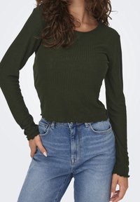 Grünes, geripptes Langarmoberteil mit rundem Ausschnitt und wellenförmigem Saum, kombiniert mit hellblauen, hoch taillierten Jeans.