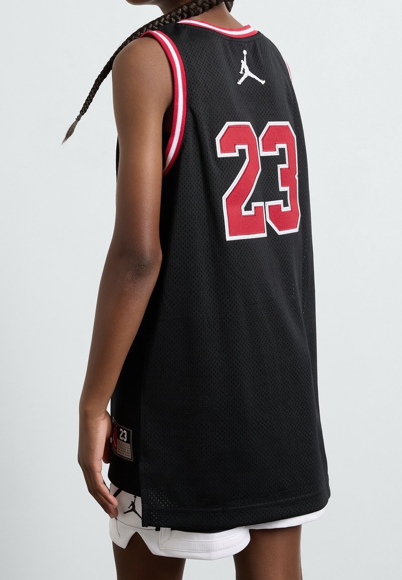 Personne portant un maillot de basket noir avec des bordures rouges et blanches, numéro 23, et un short blanc avec une coiffure tressée.