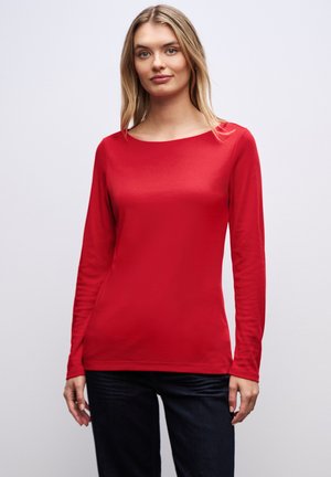 Maglia rossa a maniche lunghe realizzata in tessuto liscio, con scollo rotondo e silhouette aderente, abbinata a jeans scuri. Nessun motivo o dettaglio particolare.