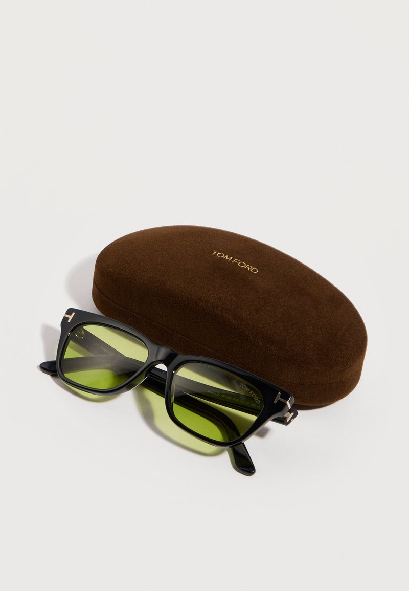 Lunettes de soleil à monture noire avec verres verts posées devant un étui rigide brun de marque Tom Ford sur une surface blanche.