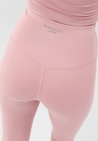 Leggings taille haute rose en matériau extensible, avec des coutures plates et une étiquette logo près de la taille pour le branding.