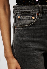 Κοντινό πλάνο ενός ατόμου που φορά ψηλόμεσα ξεθωριασμένα μαύρα τζιν με χάλκινα καρφιά και μαύρο τοπ με πολύχρωμες παγιέτες.