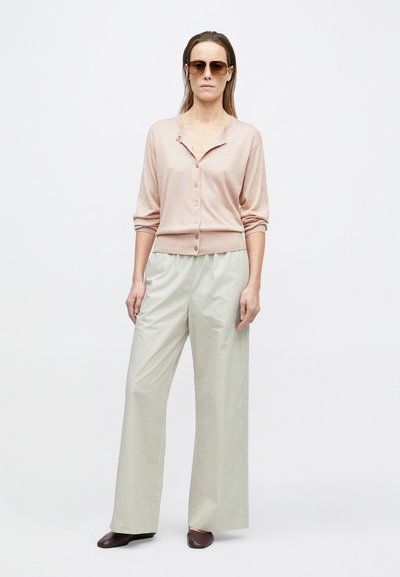 Frau trägt hellrosa Strickjacke mit Knöpfen, beige weite Hosen, dunkle flache Schuhe und Sonnenbrille, steht vor einem schlichten weißen Hintergrund.