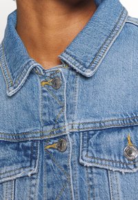 Světle modrá džínová bunda s výrazným límcem, žlutým šitím a stříbrnými knoflíky. Detailní záběr zdůrazňující strukturu tkaniny a detaily.