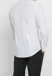 Camicia bianca a maniche lunghe con colletto classico, tessuto morbido e orlo curvo. Abbinata a pantaloni scuri, presenta un design semplice e pulito.