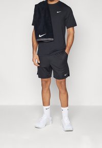 Svart Nike t-shirt och shorts, mjukt tyg, avslappnad passform. Bärande vita Nike-sneakers och strumpor. Handduk hängande över axeln, också svart.