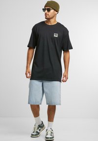 Black cotton t-shirt with short sleeves and green logo patch, paired with light blue denim shorts and green and white sneakers.