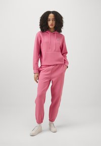 Felpa rosa con cappuccio, corde di regolazione e tasca frontale, abbinata a pantaloni jogger coordinati. Tessuto testurizzato, vestibilità rilassata e polsini elasticizzati. Sneakers bianche.