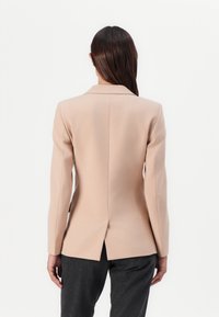 Svetle béžový tailor-fit blazer s klopový límcem, dlouhými rukávy a rozparkem vzadu, kombinovaný s tmavě šedými dobře střiženými kalhotami. Hedvábně hladký povrch.