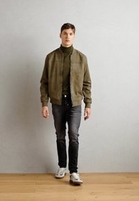 Maslinasta suede bomber jakna, visoki ovratnik, dvije džepove; nosi se preko tamnozelene rolke, uparena sa uskim tamnim trapericama i bijelim tenisicama.