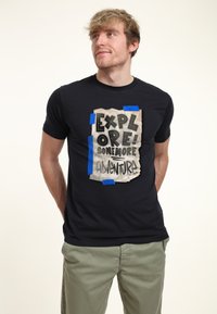 Černé tričko s grafickým designem potrhaného a přelepeného papíru s nápisem "PROZKOUMEJ! JEŠTĚ VÍCE DOBRODRUŽSTVÍ!" v tučných, hravých písmenech.