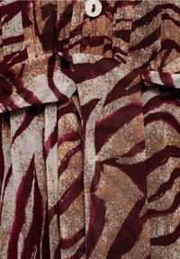 Gros plan sur un tissu avec des motifs abstraits marron, blanc et bordeaux, comportant un bouton et un détail de ceinture nouée.