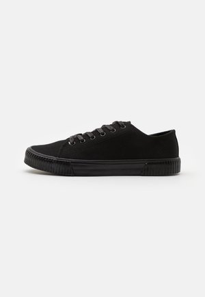 Zapatilla deportiva baja de lona negra con cordones en la parte delantera y suela de goma texturizada sobre fondo blanco.