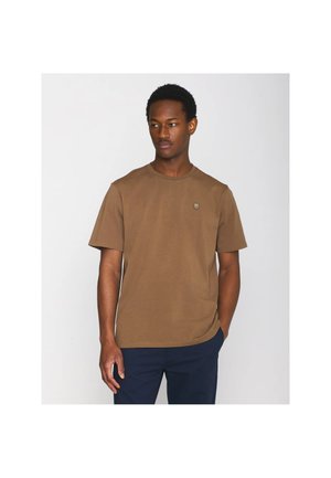 Man draagt een bruin T-shirt met korte mouwen met een klein logo op de borst en donkere broek, staand met één hand in de zak tegen een witte achtergrond.