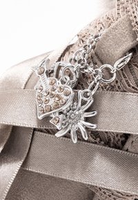 Bracelet porte-bonheur en argent avec un pendentif en forme de cœur orné de strass et un pendentif en forme de fleur, sur un fond de tissu beige texturé.