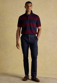 Svītrota polo krekls tumši sarkanā un jūras zilā krāsā, ar apkakli un īsām piedurknēm. Apvienots ar jūras zilajiem chinos un brūnajām mokasīnām, novietots pret bēšainu fonu.