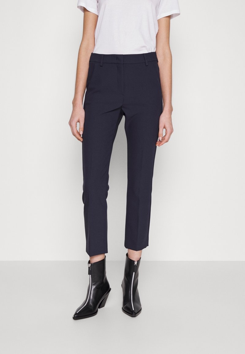 Femme portant un pantalon slim bleu marine, un haut blanc à manches courtes et des bottines noires à talons avec fermeture éclair à l'avant, debout devant un fond uni.