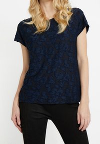 Granatowa bluzka z krótkim rękawem o teksturowanym floralnym wzorze, wykonana z lekkiego materiału; luźny krój, okrągły dekolt i podwinęte rękawy.