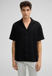 Schwarzes kurzärmliges Hemd mit einem strukturierten Zickzackmuster, Knopfverschluss und Kragen. Kombiniert mit hellen Hosen.