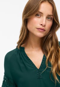 Blusa verde scuro con scollatura a V e dettagli in pizzo testurizzato al colletto. Spalle leggermente arricciate, tessuto liscio e vestibilità casual.