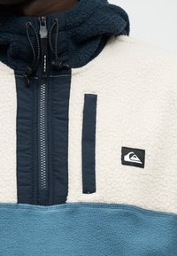 Pusaujas flīsa jaka ar tumši zilu rāvējslēdzēju, krēmkrāsas augšējo daļu, zilu apakšējo daļu, melnām regulējamām auklām un nelielu logotipa plāksteri uz krūtīm.