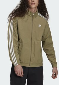 Olivgrön zip-up jacka i lätt material, med vita tre-rands accenter på ärmarna och en liten vit logotyp på bröstet.