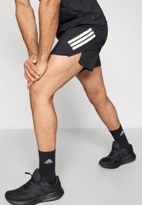 Čierne športové šortky s bielymi pruhmi na bokoch, doplnené čiernymi ponožkami s logom a čiernymi športovými topánkami s textúrovanou podrážkou.