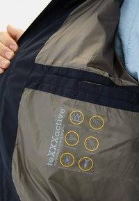 Waterproof jacket interior with recycle emblem, beige lining, and black trim. Features icons for waterproof, breathable, windproof, and taped seams.